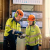 A male and a female recycling expert, both wearing hardhat, hearing protection, protective glasses and reflective work clothes, are discussing a document held by the male expert.